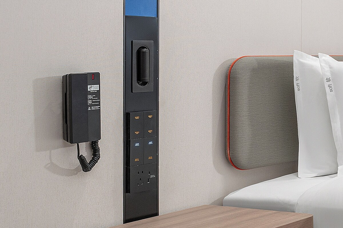A hotel room with a built-in telephone, a control panel, and a bed with white bedding and a grey headboard.