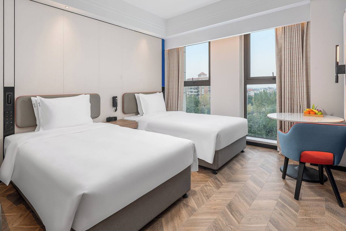 A hotel room with two beds, a desk, a chair, and a large window with a city view.