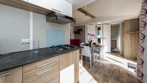 A modern kitchen with a gas stove, wooden cabinets, and a dining area with a table and chairs. - ZEcamping 4 étoiles animé parc aquatique locatif TV 30m2 4 pers (Fouesnant)