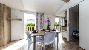 A dining area with a round table, white chairs, and a view of the outdoors. - ZEcamping 4 étoiles animé parc aquatique locatif TV 30m2 4 pers (Fouesnant)