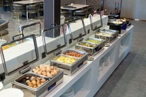 Een buffet met diverse etenswaren in metalen bakken, gerangschikt op een toonbank met een glazen scheiding.