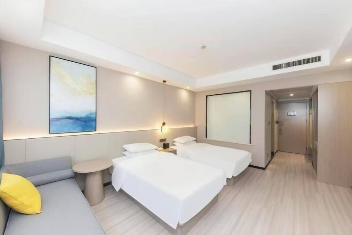 Una habitación de hotel moderna con dos camas individuales, un sofá, una mesita pequeña y un ventanal grande.
