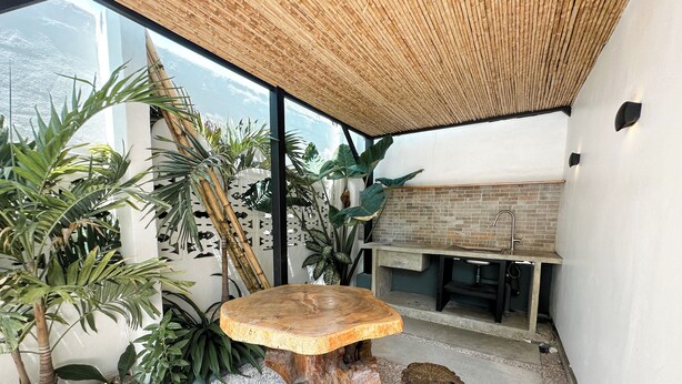 A modern outdoor dining area with a round wooden table and stools, surrounded by lush greenery and a view of the sea.