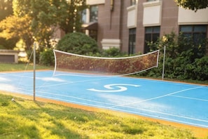 Una cancha deportiva con una red, rodeada de césped y árboles, con un edificio al fondo.