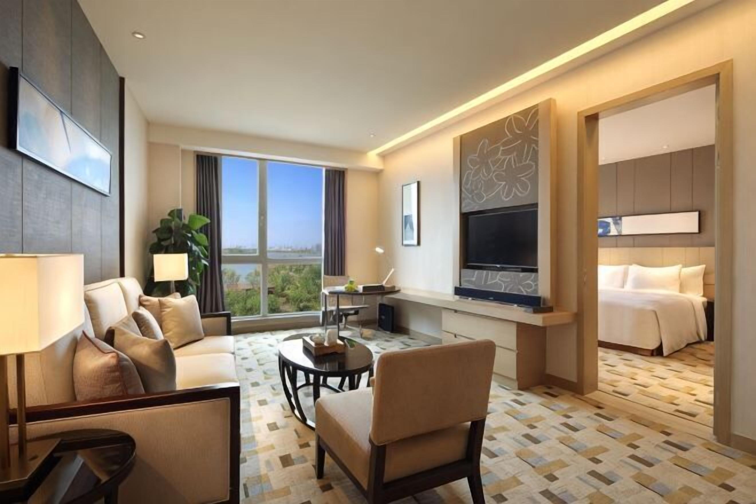 A hotel room with a sofa, armchair, coffee table, TV, and a balcony with a view.