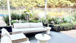 A deck with a white sofa, wicker chairs, and a round table, surrounded by potted plants and a wooden fence. - SERENITY STAYS: Best price for value - 5 min from beach, cafes and racecourse (Mount Martha)
