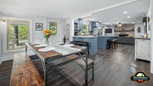 A modern kitchen with a central island, a dining area with a wooden table set for four, and a view of the outdoors through large windows. - Pet Friendly, Short walk to Lake Monroe (Nashville)