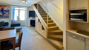 A modern apartment with a staircase leading to a loft bedroom, a dining area with a table and chairs, a kitchen with a microwave, and a living room with a sofa and a TV.