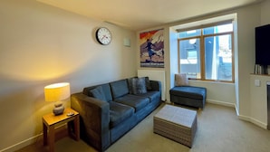 A living room with a sofa, a small table with a lamp, a clock on the wall, a poster, and a window with a view.
