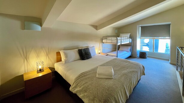 A bedroom with a bunk bed, a single bed, a bedside table with a lamp, and a window with a view.