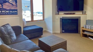A living room with a large flat-screen TV, a fireplace, a sofa, and a window with a view of parked cars outside.