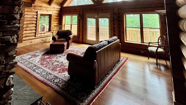 L’intérieur d’une cabane en rondins, avec un plafond haut, des poutres en bois, une cheminée, un canapé et une grande fenêtre donnant sur une terrasse.