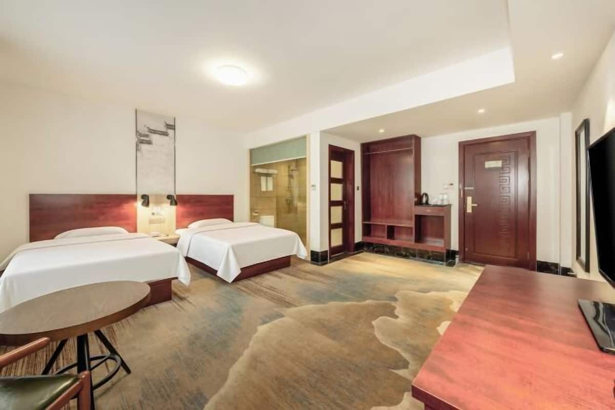 Habitación de hotel con dos camas, un escritorio, una silla, un televisor, un espejo y un baño visible a través de una puerta abierta.