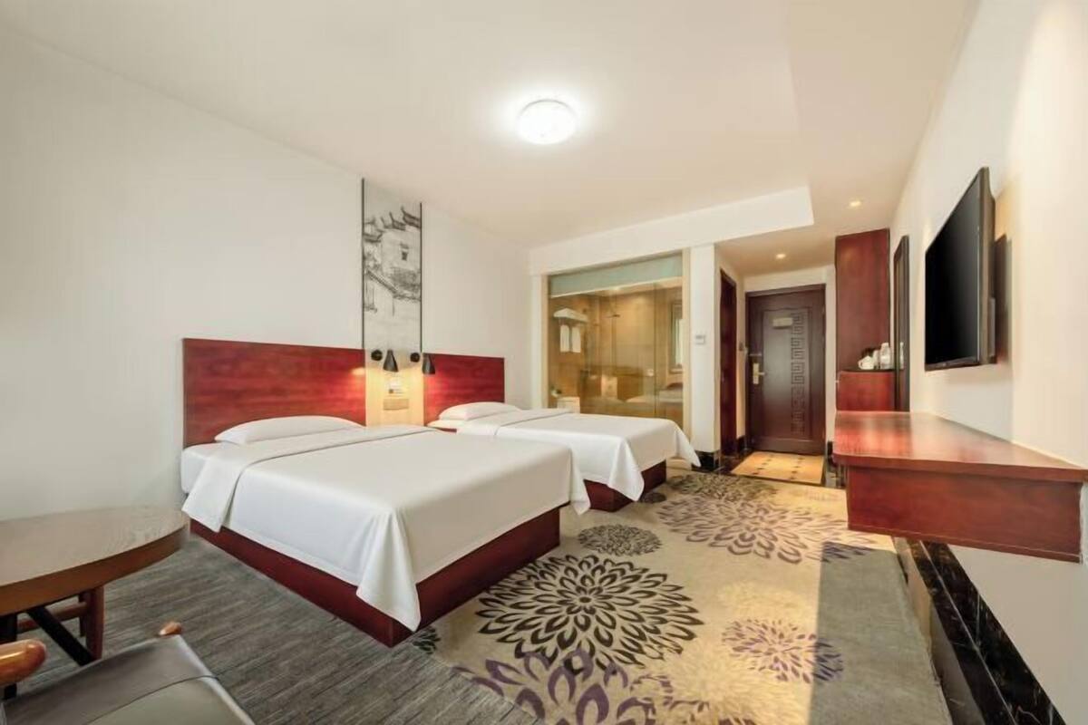 Habitación de hotel con dos camas, un escritorio, un televisor y un baño visible a través de una puerta de cristal.