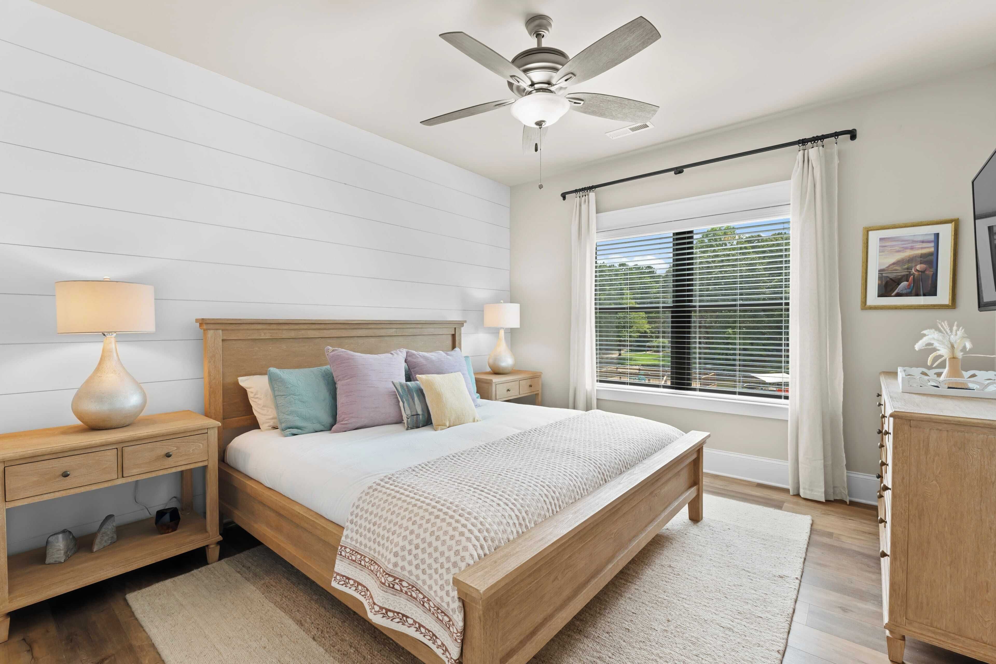 A bedroom with a wooden bed, nightstands, a ceiling fan, a window with blinds, and a night lamp.