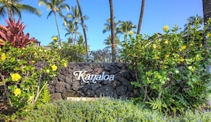 A stone wall with the name "Kanaloa" in the center, surrounded by lush greenery and palm trees. - Aloha Condos, Kanaloa at Kona, Condo 3103, Oceanfront, AC (Kailua Kona)