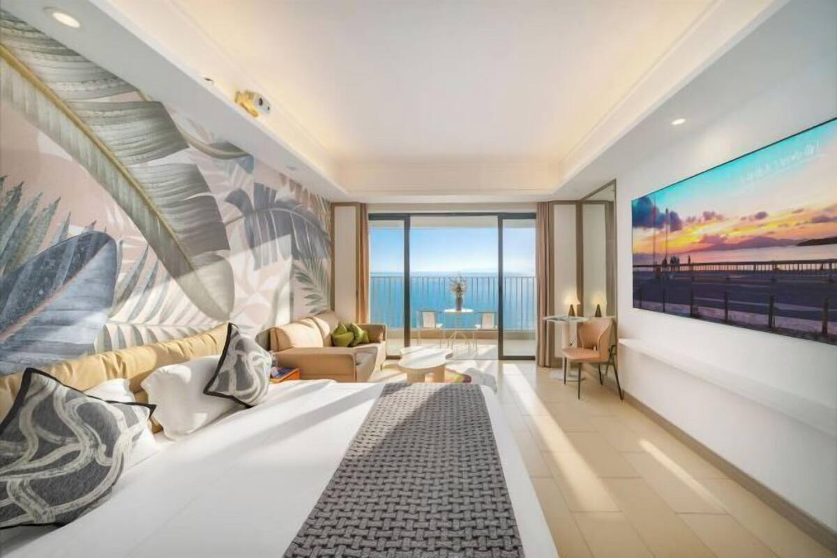 A modern hotel room with a large bed, a sofa, a dining area, and a balcony with a view of the sea.