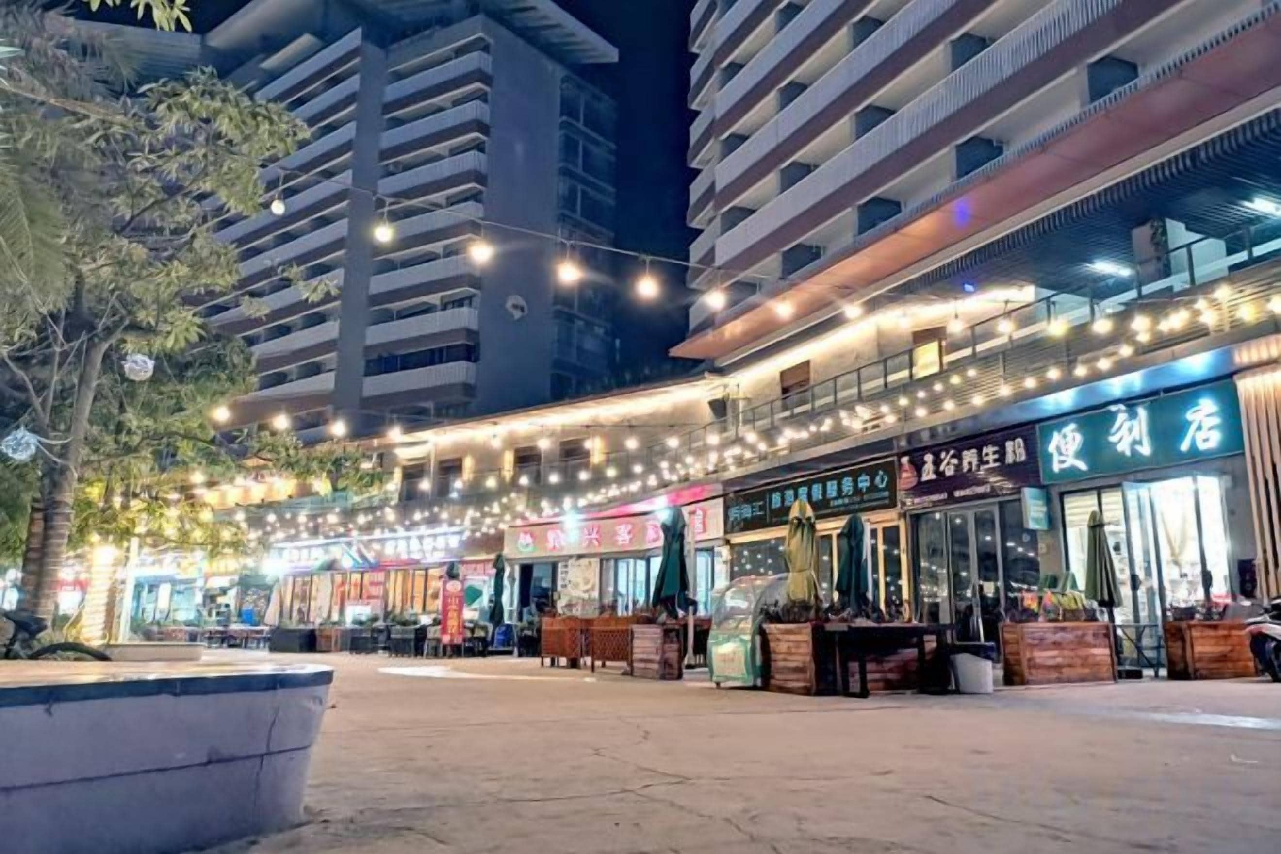A commercial area with multiple storefronts, including a pharmacy and a restaurant, under a string of lights.