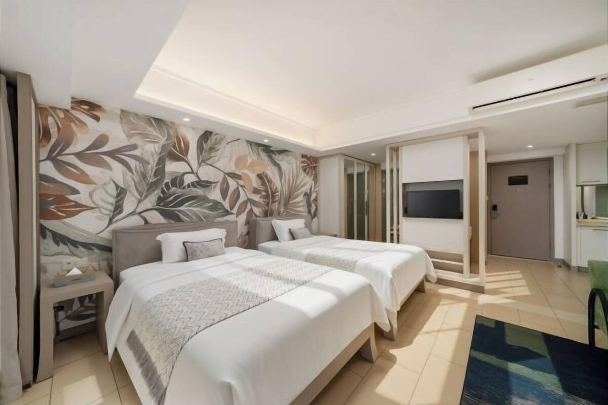 A modern hotel room with two beds, a large window, a flat-screen TV, and a decorative leaf-patterned wall.