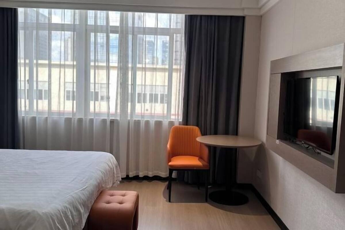 Ein Hotelzimmer mit einem Bett, einem kleinen runden Tisch, einem Stuhl, einer Bank und einem großen Fenster mit Vorhängen.