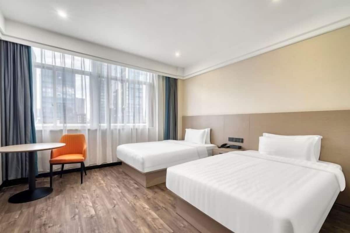 Ein Hotelzimmer mit zwei Betten, einem kleinen Tisch, einem orangefarbenen Sessel und einem großen Fenster mit Vorhängen.