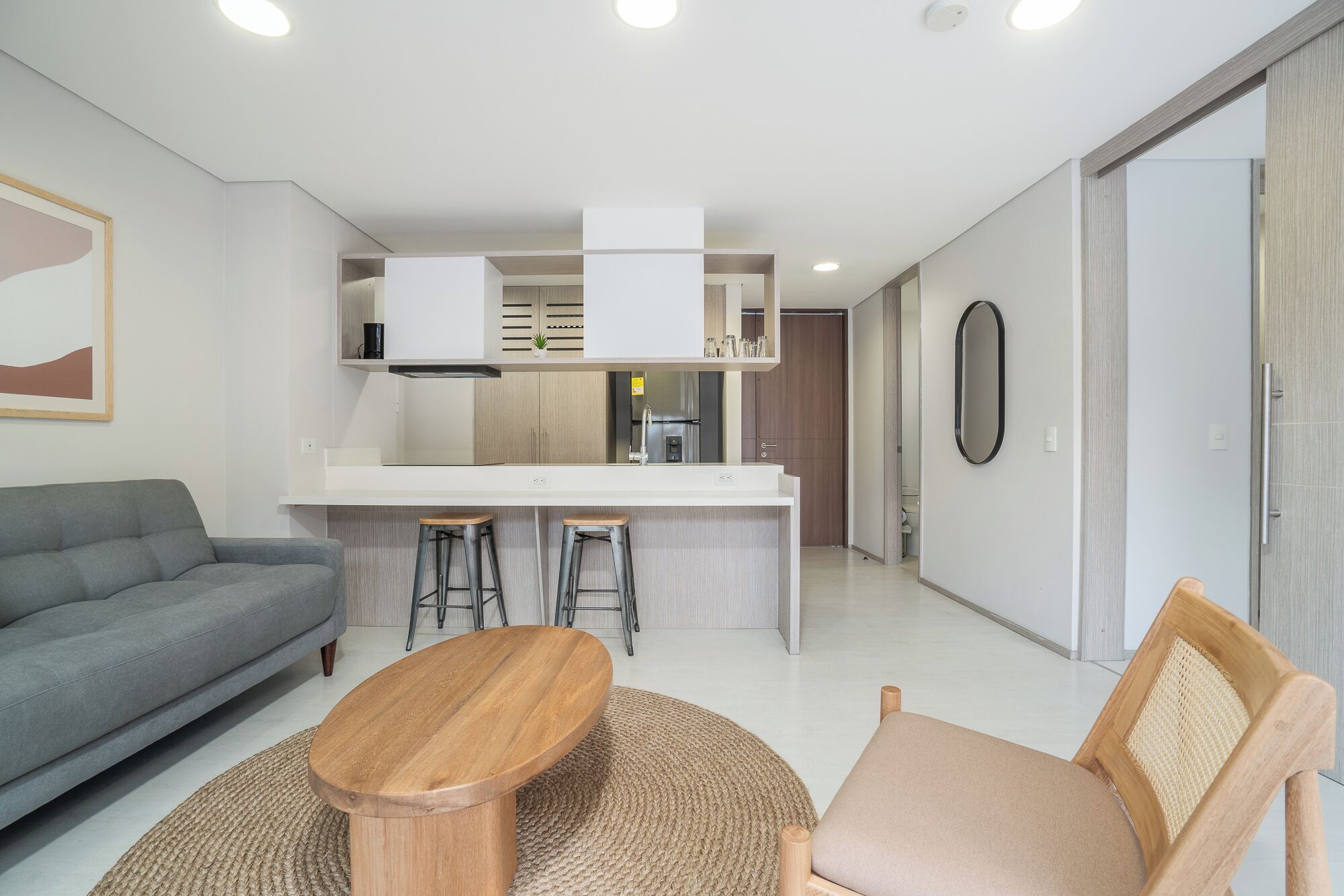 A modern living room with a grey sofa, a wooden coffee table, and a wicker chair. A kitchen area with a white countertop and stools is visible in the background.