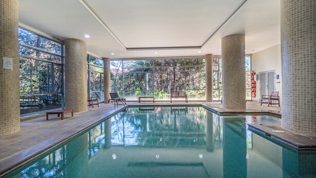 Una piscina interior amb parets de vidre transparent, seients de fusta i columnes de rajola.