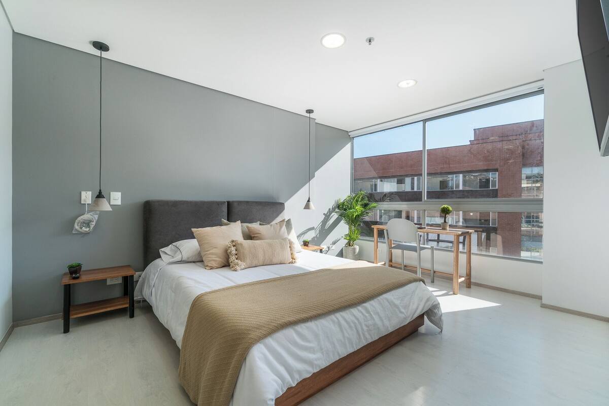 A modern bedroom with a large bed, bedside tables, a small desk, and a large window offering a view of the city.