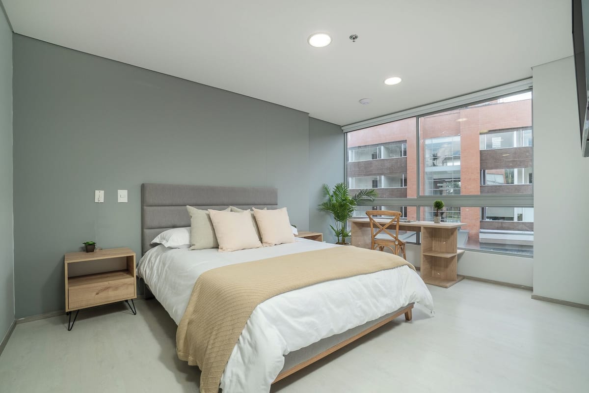 A modern bedroom with a large bed, bedside tables, a desk, and a window with a view of buildings.