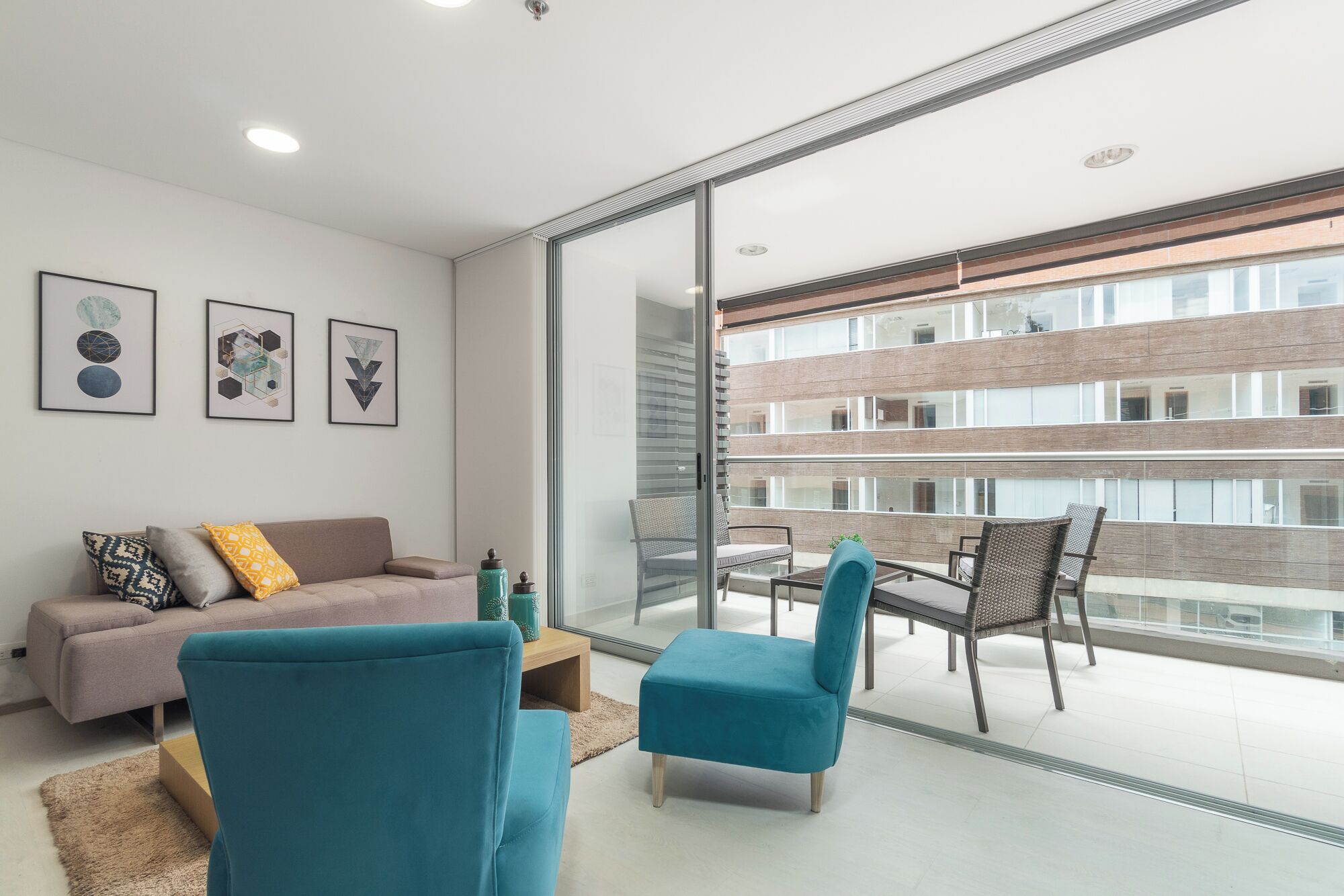 Una sala moderna con un sofá, un sillón turquesa y una mesa de vidrio. Hay obras de arte enmarcadas en la pared y un balcón con vistas a un edificio.