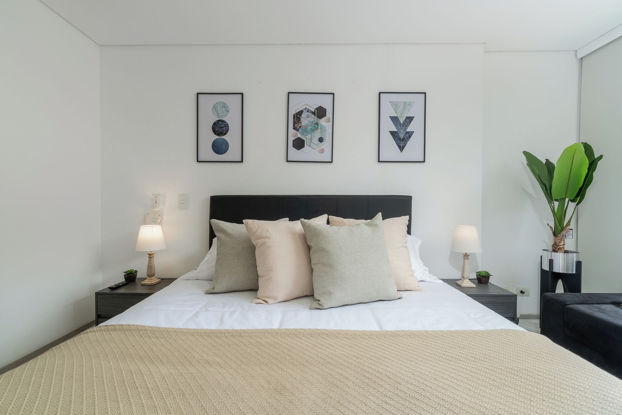Un dormitorio ordenado con una cama grande, dos mesitas de noche con lámparas y tres cuadros en la pared.