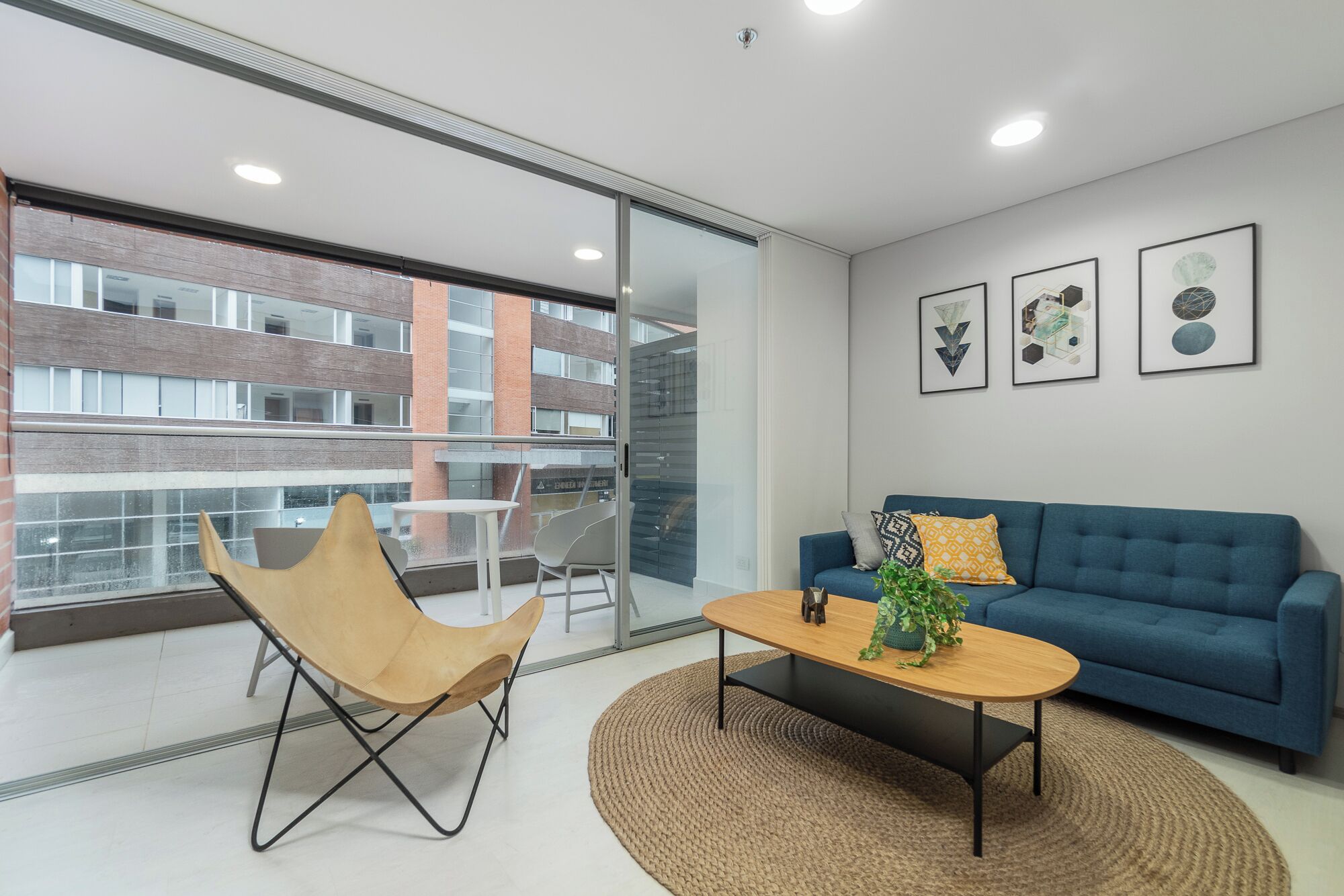 Una sala de estar moderna con un sofá azul, una mesa de centro de madera y un sillón color arena. Hay obras de arte enmarcadas en la pared y una vista de un edificio a través de una puerta de vidrio.