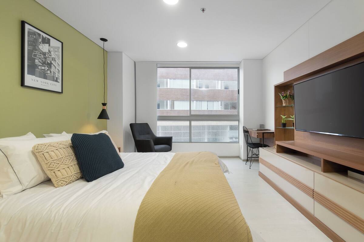A modern hotel room with a large bed, a desk, a chair, a TV, and a window with a view of buildings.