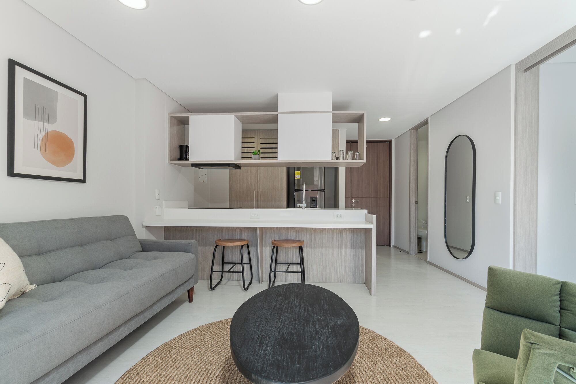 A modern living room with a grey sofa, a round black coffee table, and a green armchair. A kitchen area is visible with a white countertop, a refrigerator, and a sink.