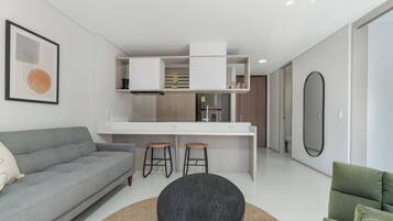 A modern living room with a grey sofa, a round black coffee table, and a green armchair. A kitchen area is visible with a white countertop, a refrigerator, and a sink.