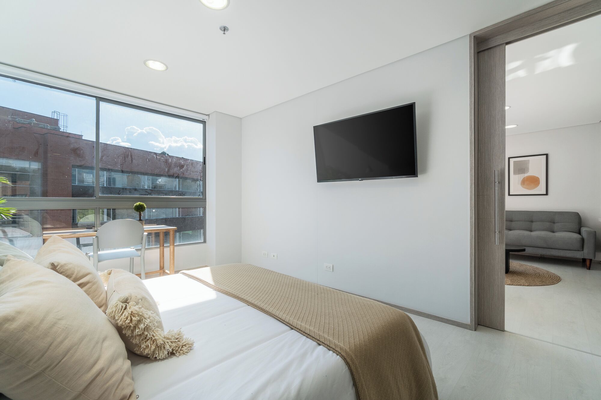Une chambre moderne avec une grande fenêtre, un lit avec une literie beige, un téléviseur fixé au mur et un canapé gris dans le salon attenant.