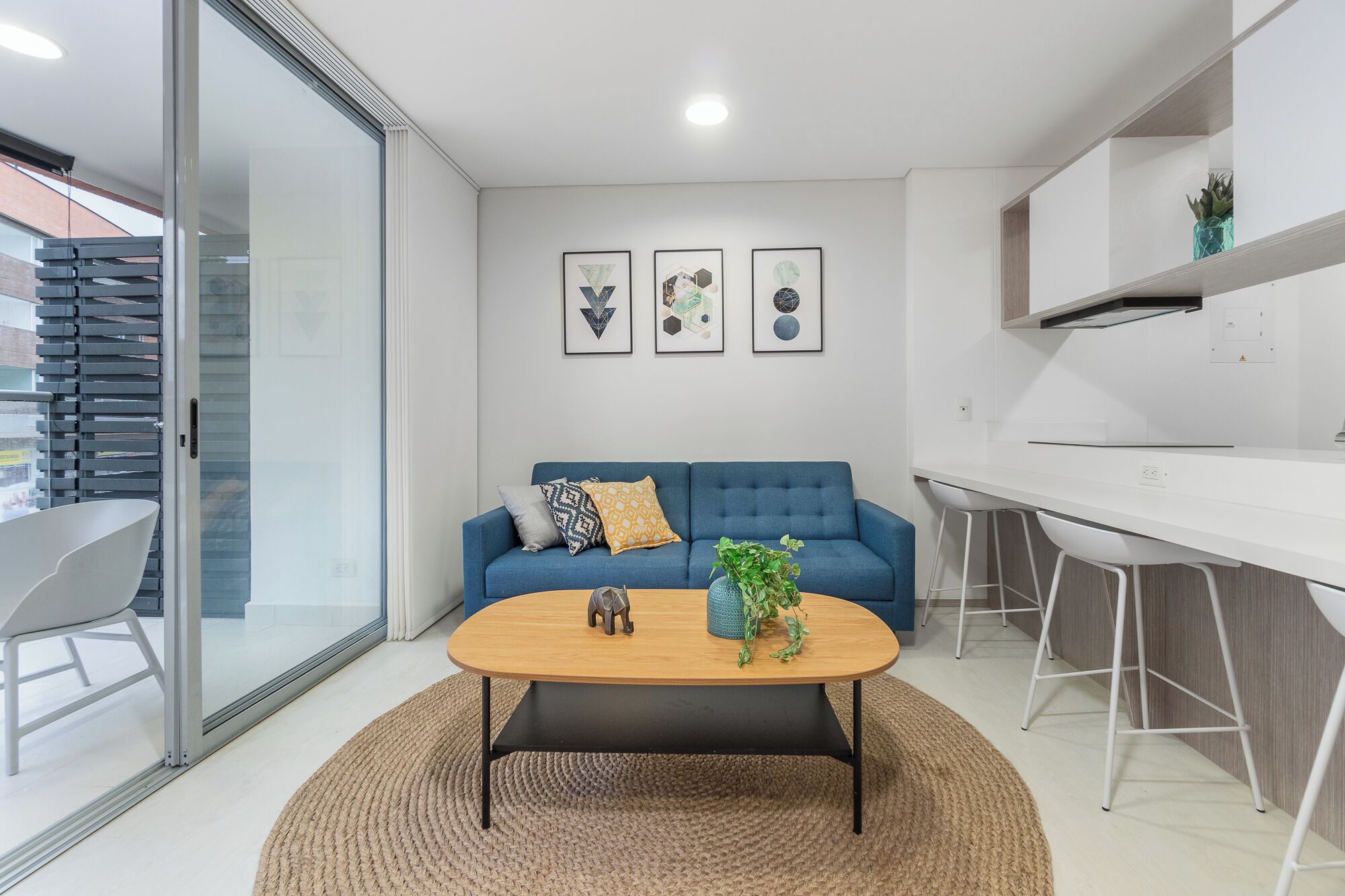 A modern living space with a blue sofa, a wooden coffee table, and a kitchen area with white cabinets and bar stools.