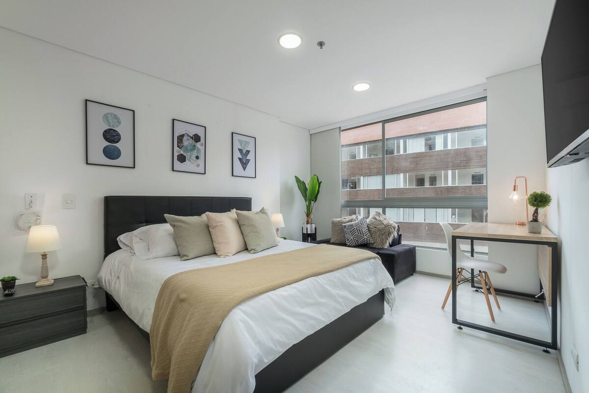 A modern bedroom with a large bed, bedside tables, a desk, and a window with a view of a brick building.