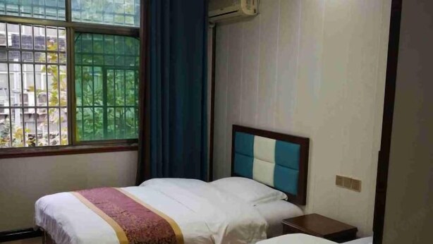 A hotel room with two beds, a window with bars, a bedside table, and an air conditioning unit. - Enshi Xueyuan Hotel (Enshi City)