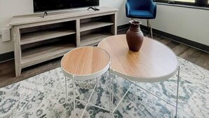 A modern room with a flat-screen TV on a wooden stand, a blue chair, and a small round table with a vase. - Sleepover | Superior 1BD/1BA - Des Moines (West Des Moines)