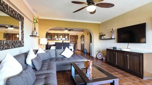 A living room with a sectional sofa, a coffee table, and a TV. There is a kitchen in the background with cabinets and a refrigerator. - A-505 Enjoy Your Vacation In This Charming Condominium! (Cabo San Lucas)
