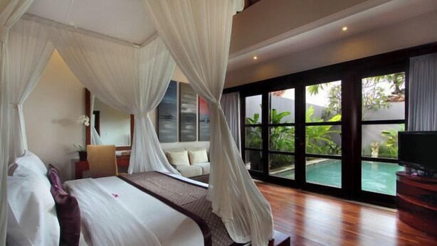A bedroom with a canopy bed, a view of an outdoor swimming pool, and a television on a wooden stand. - Aria Exclusive Villas & Spa (Bali)