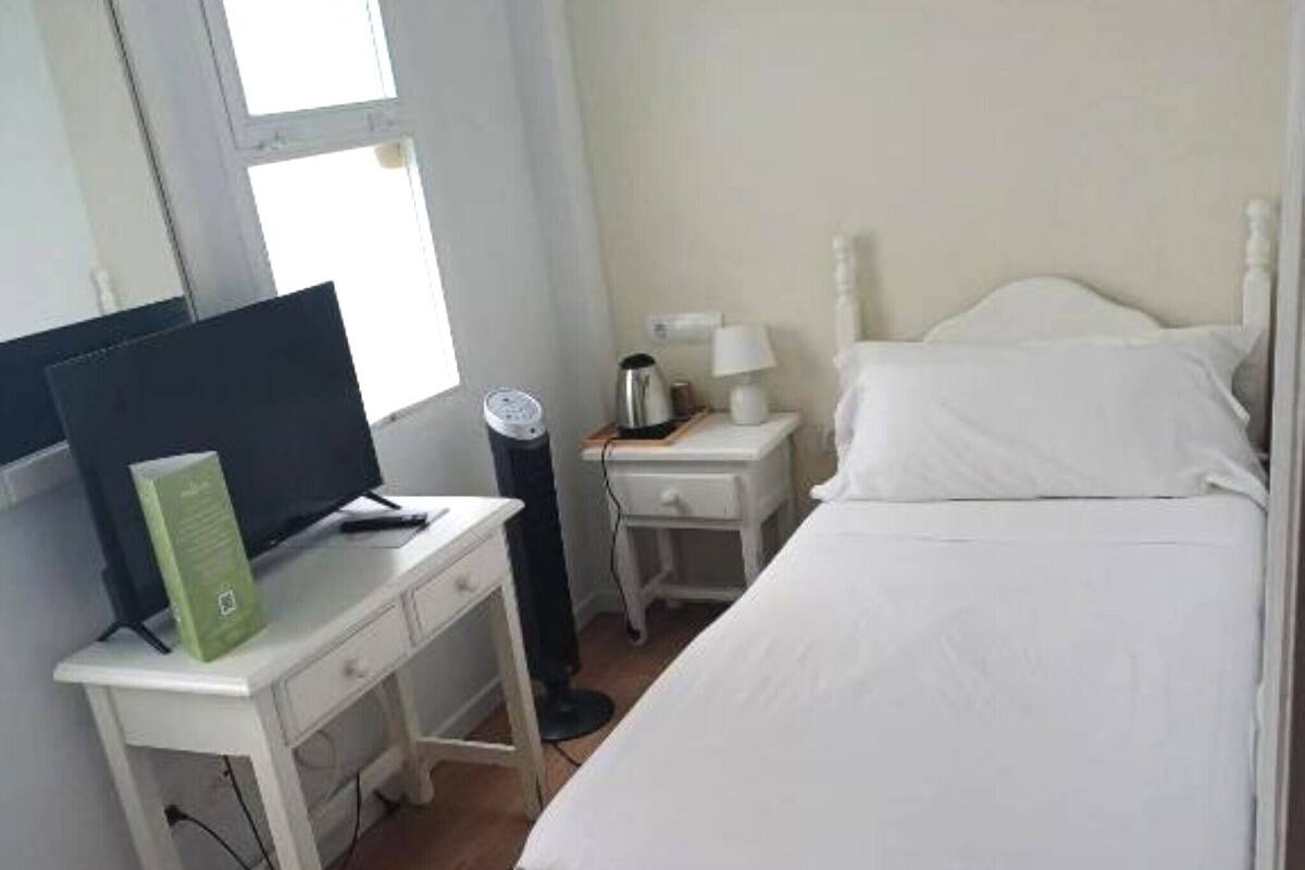 A bedroom with a bed, a desk with a computer, a nightstand with a lamp, and a window.