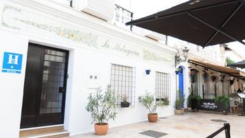 Un edificio con un letrero que dice "La Malaguena" y una señal azul con la letra "H" que indica un hotel. Hay plantas en macetas y una zona de estar al aire libre cubierta.
