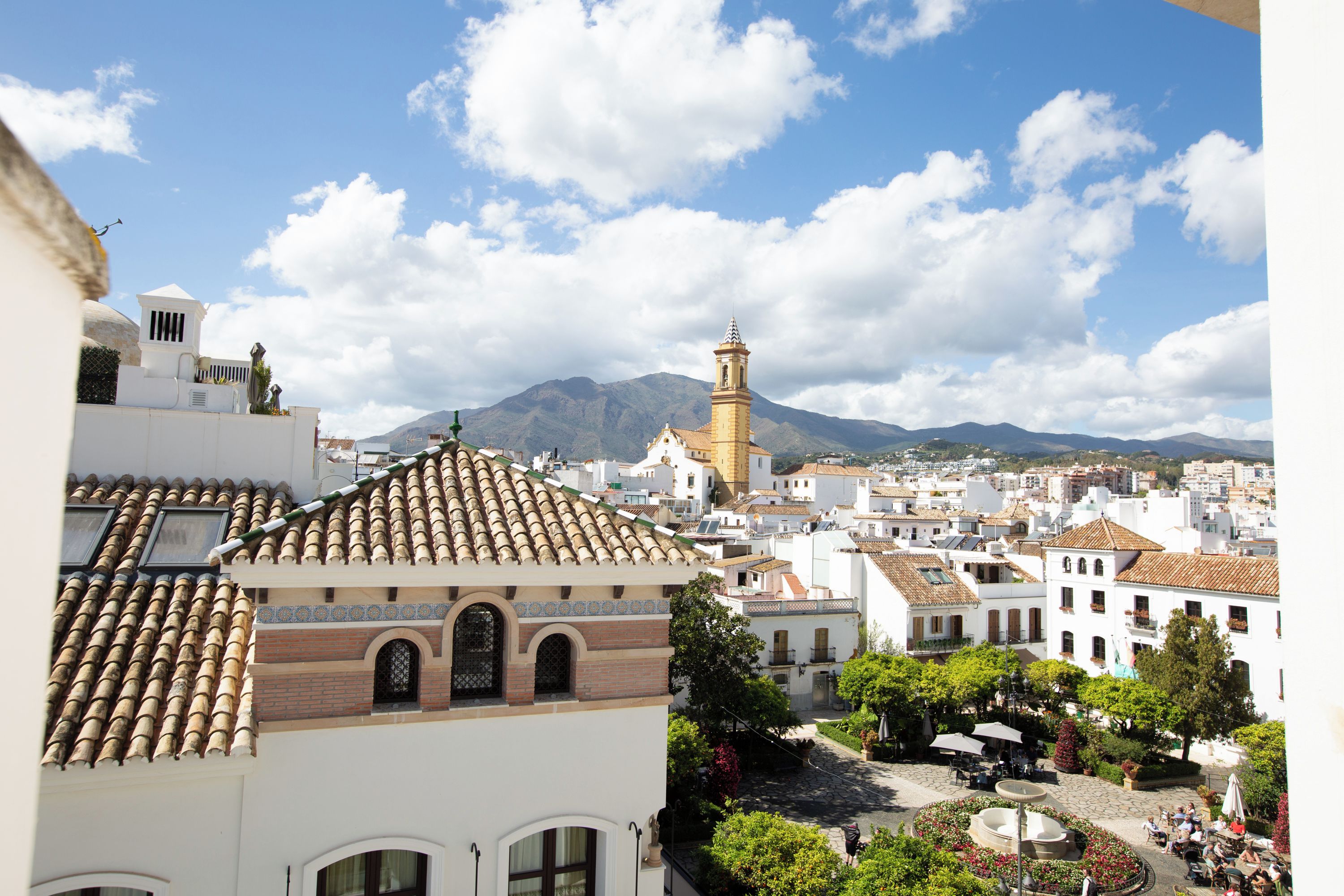 Una plaza de pueblo con una fuente, rodeada de edificios de arquitectura tradicional y una montaña al fondo.