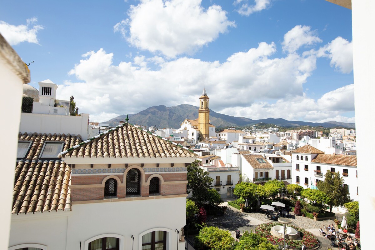 A town square with a fountain, surrounded by buildings with traditional architecture and a mountain in the background.