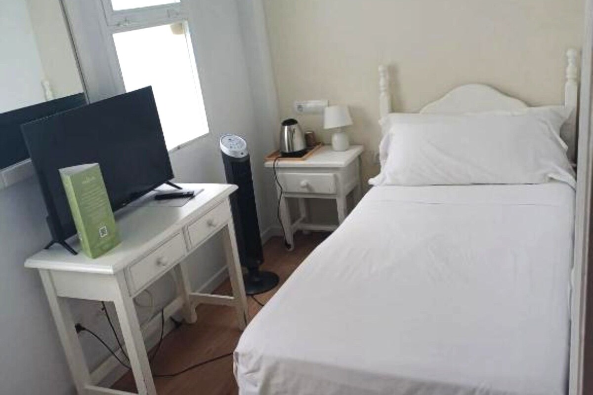 A bedroom with a bed, a desk with a computer, a nightstand with a lamp, and a window.