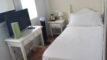 Un dormitorio con cama, escritorio con computadora, mesita de noche con lámpara y ventana.