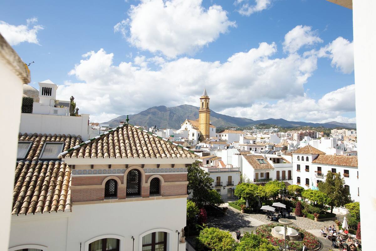 A town square with a fountain, surrounded by buildings with tiled roofs and a prominent tower.
