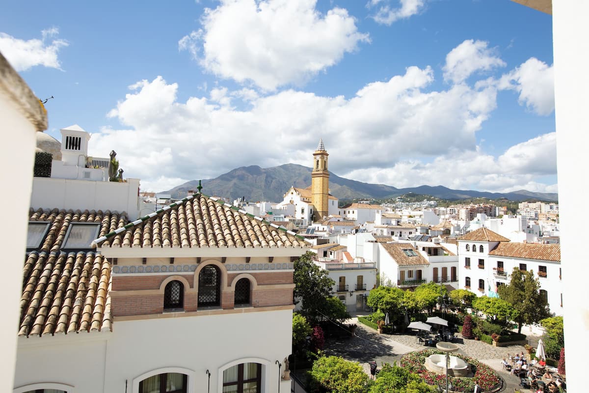 Una plaza de pueblo con edificios, una fuente y una montaña al fondo.